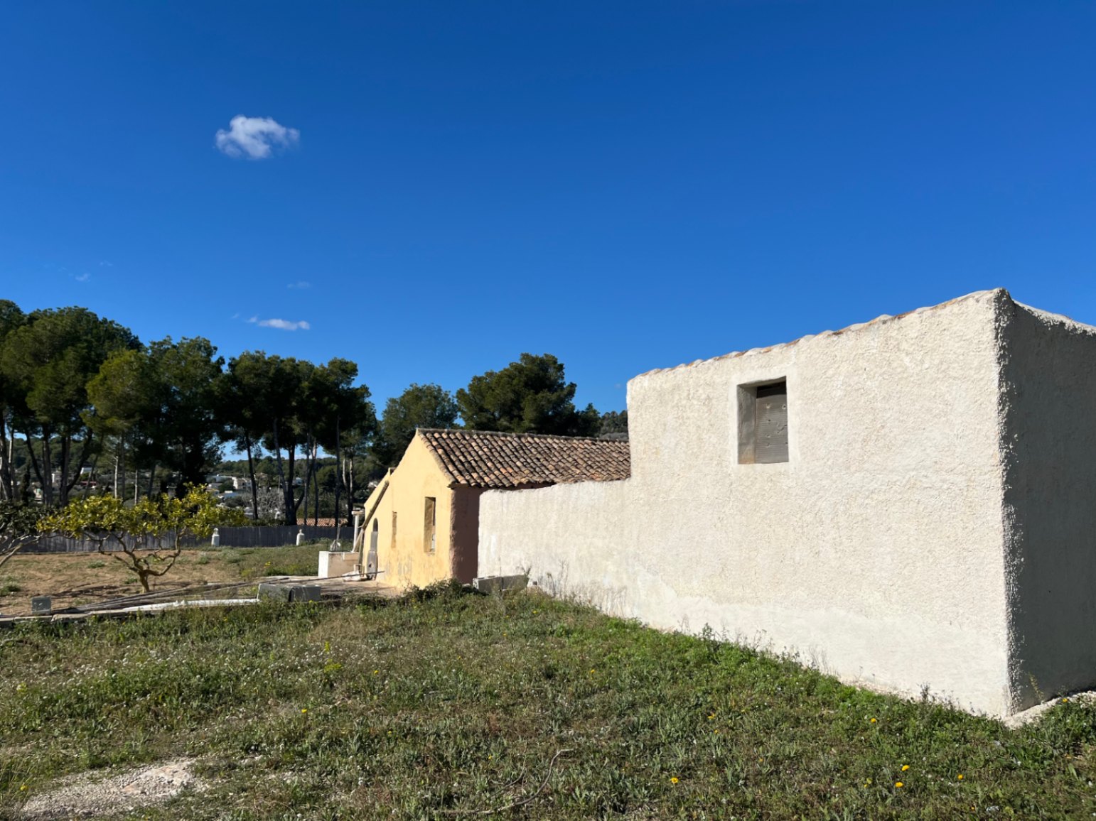 Vue extérieure de la finca traditionnelle avec murs en crépi blanc et ocre, toiture en tuiles romaines et terrain naturel sous un ciel dégagé.