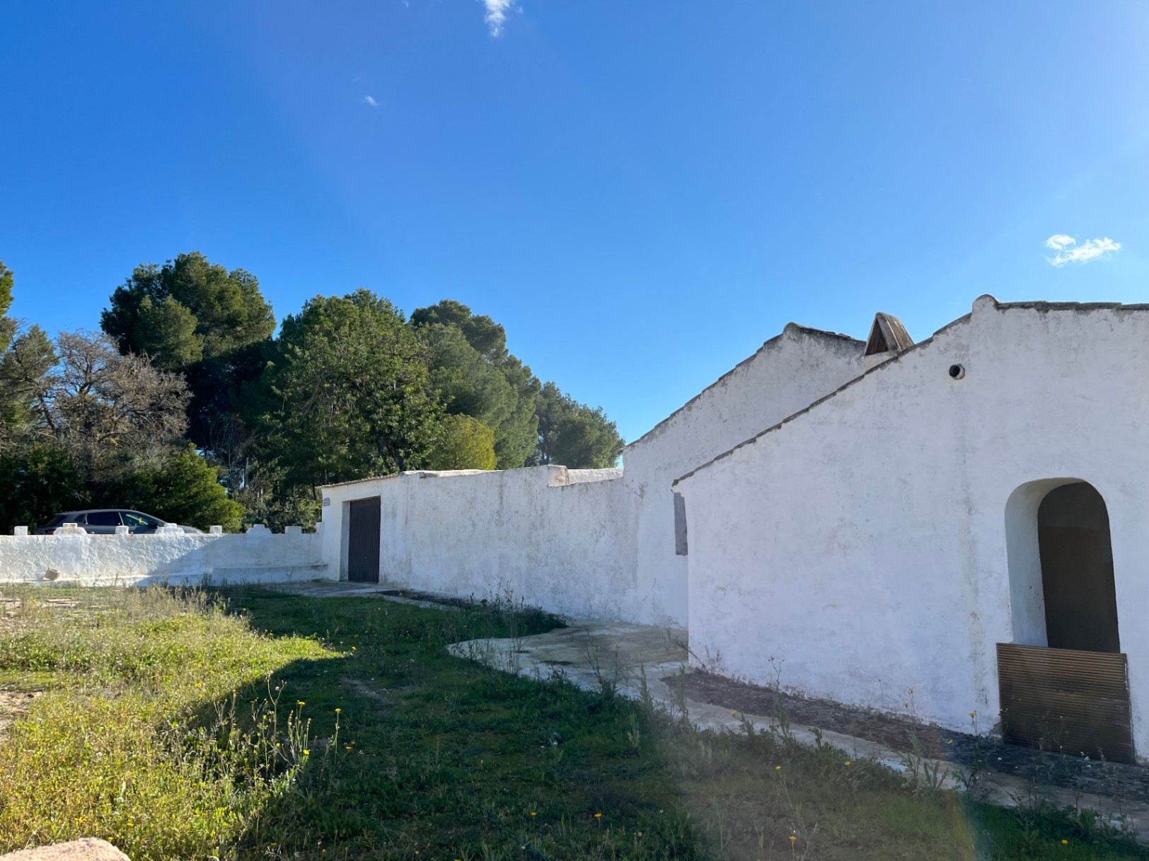 Vue extérieure d'une finca traditionnelle à Jávea, présentant des murs blanchis à la chaux, une entrée voûtée et un terrain naturel bordé de pins.
