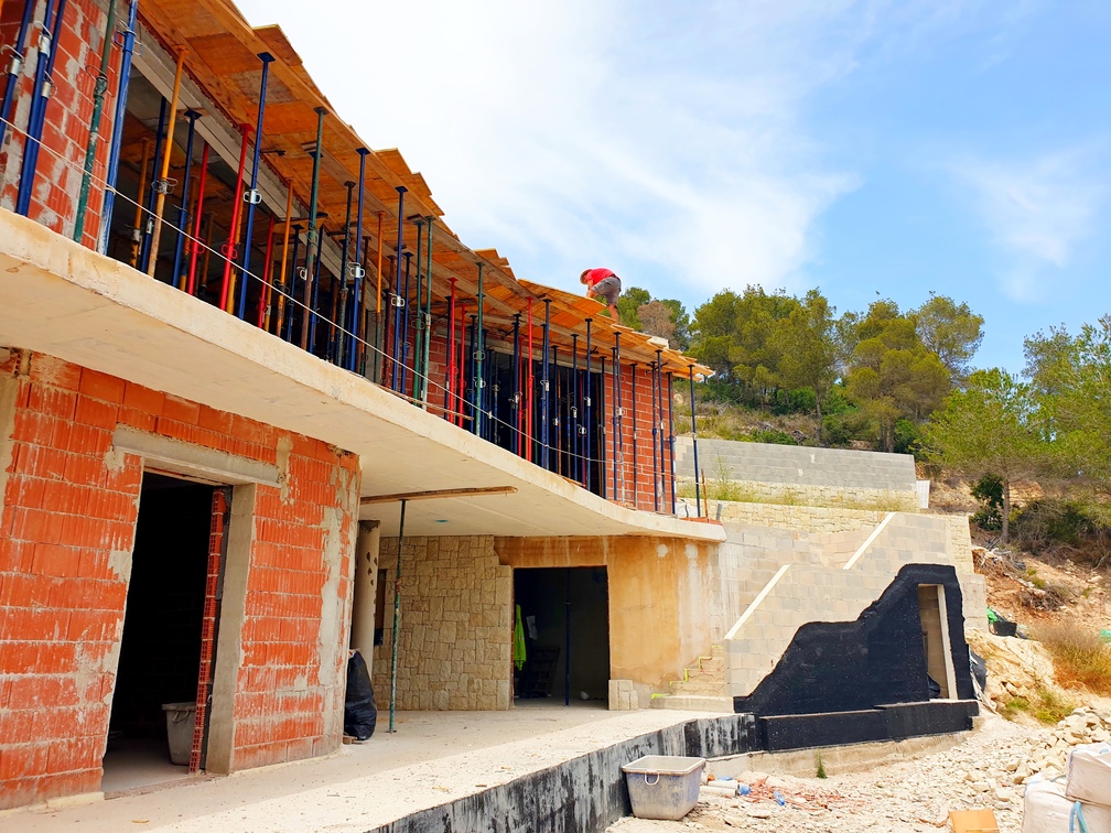 Villa moderne en construction présentant des murs en briques rouges, des dalles en béton et des étais. La structure inclut un mur en pierre et un escalier extérieur.