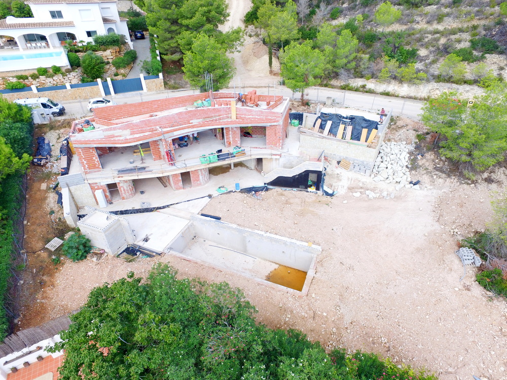 Vue aérienne d'une villa en construction présentant une maçonnerie en briques rouges, des dalles en béton et une future piscine sur un terrain en pente.