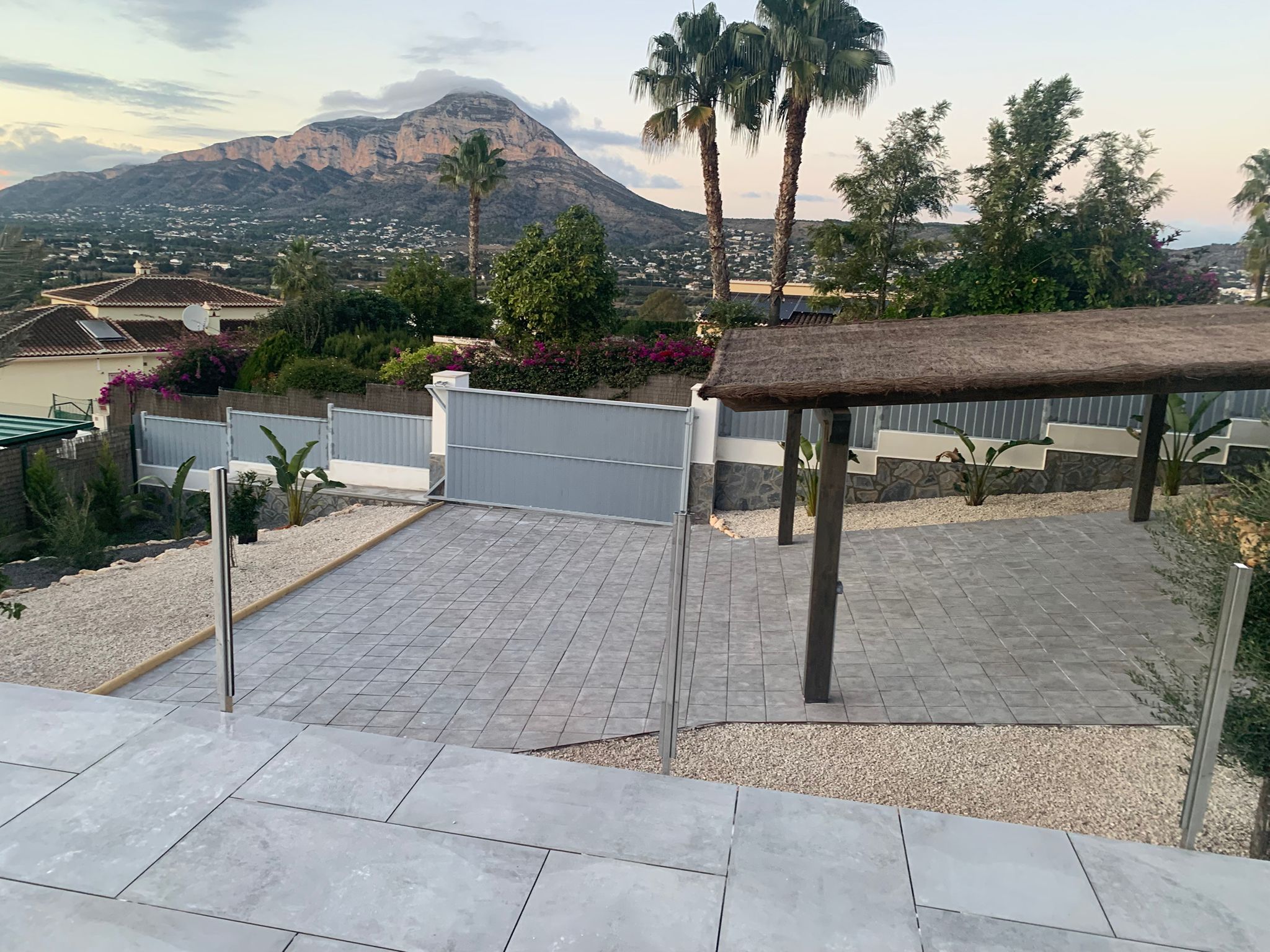 Entrée extérieure pavée de dalles grises, pergola en bois avec toit en chaume et vue dégagée sur la montagne environnante au crépuscule.