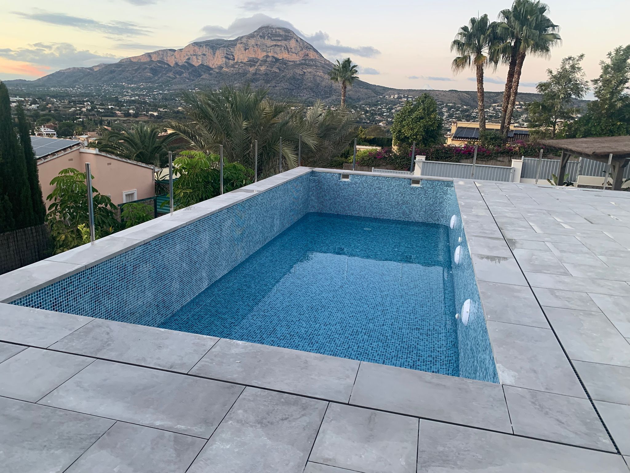 Piscine rectangulaire surélevée en mosaïque bleue avec terrasse en dalles grises, garde-corps en verre et vue panoramique sur le mont Montgó.