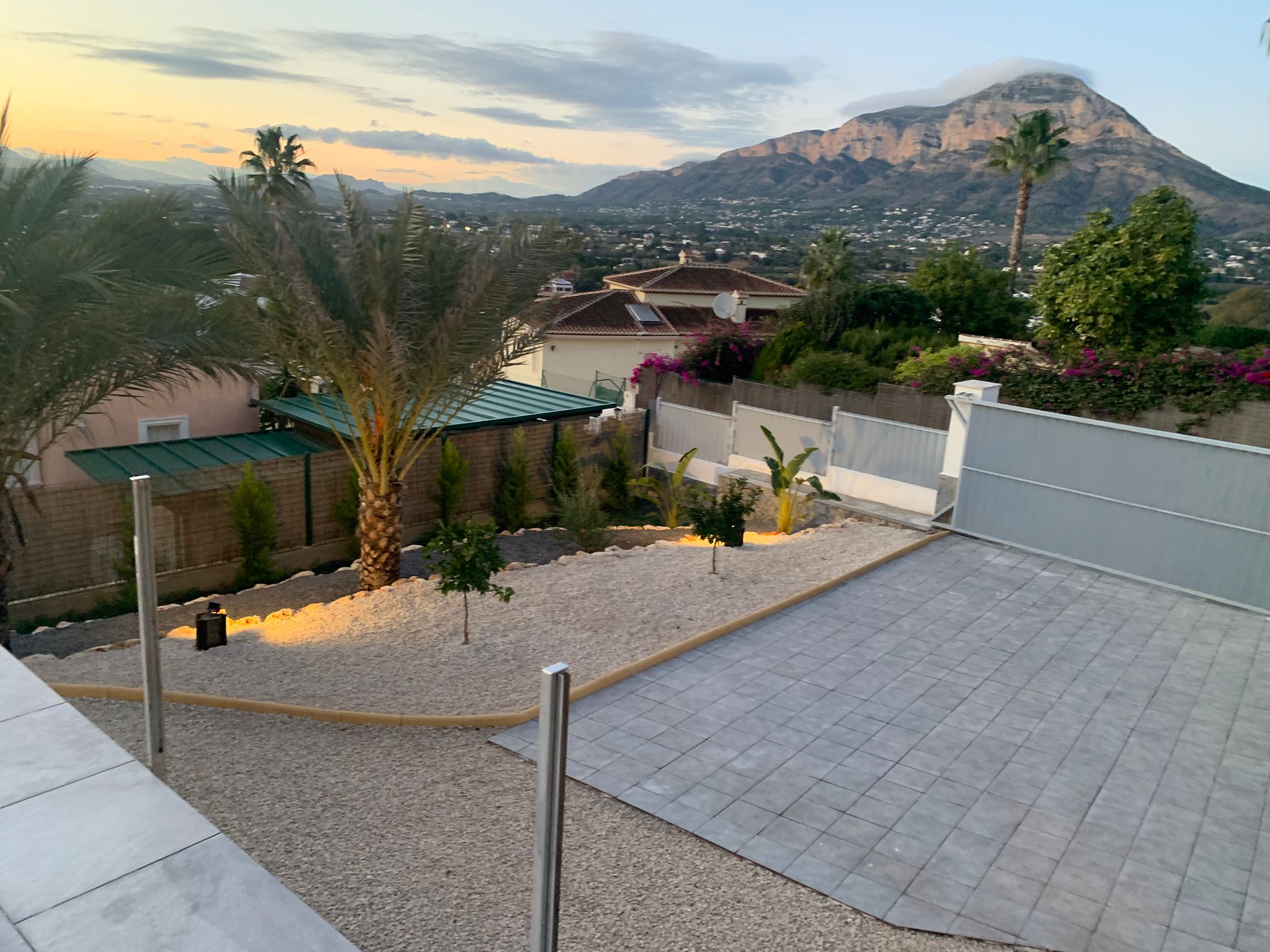 Vue surélevée d'un jardin paysager avec gravier, palmiers et allée pavée, offrant une vue panoramique sur les montagnes au coucher du soleil.