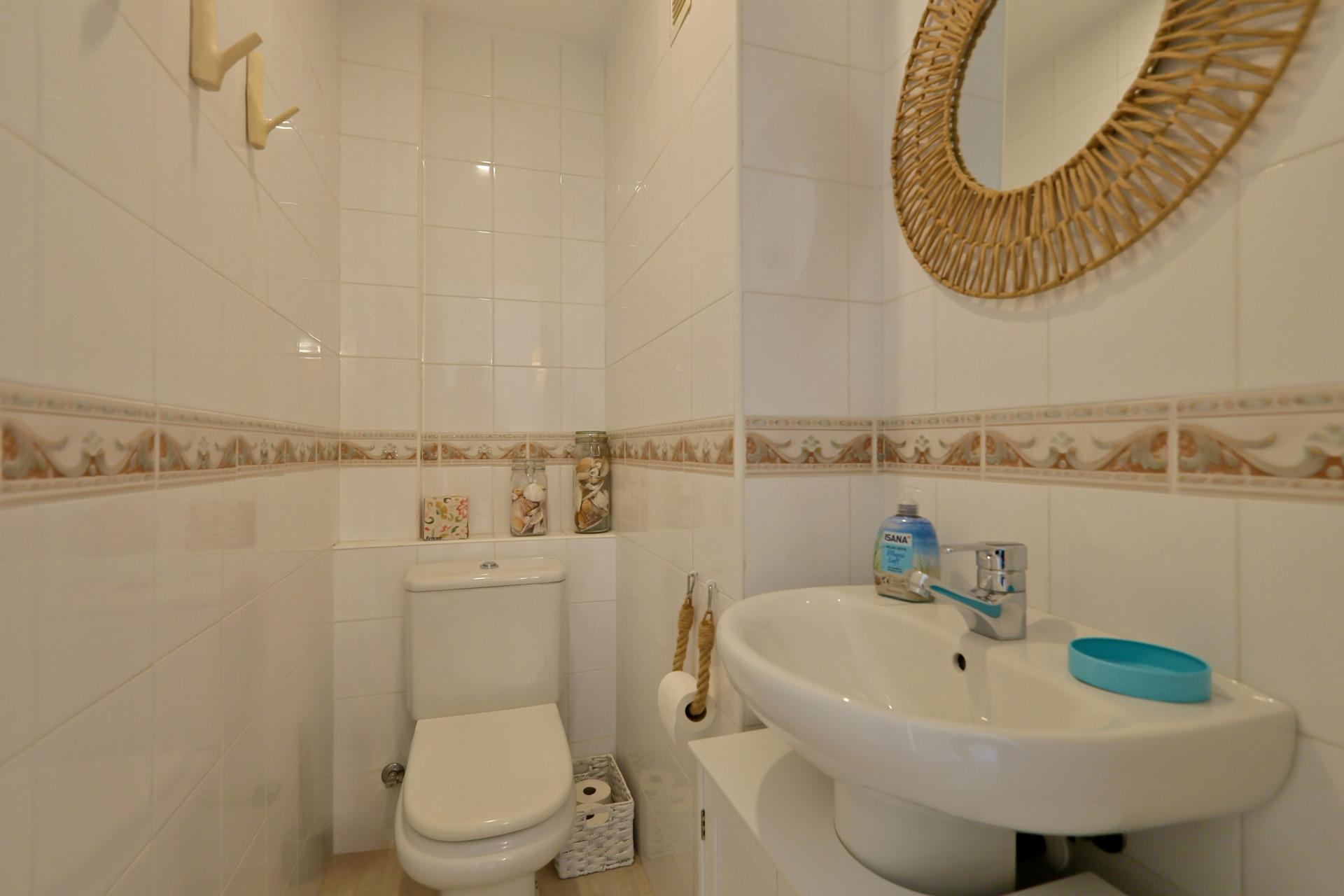 Cabinet de toilette fonctionnel avec murs carrelés blancs, frise ornementale, lavabo sur colonne et miroir circulaire en osier.