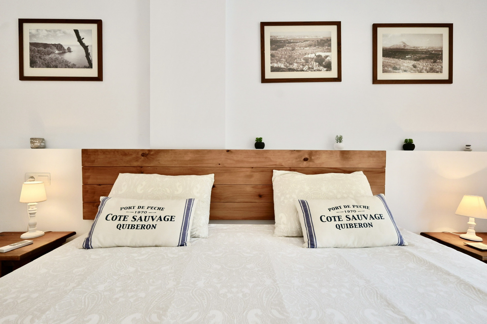 Chambre avec tête de lit en bois rustique, coussins à thème maritime et trois photographies de paysages. Tables de chevet symétriques avec lampes blanches classiques.