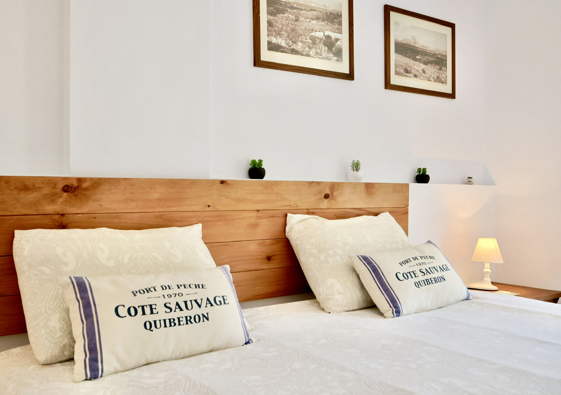 Détail de chambre avec tête de lit en planches de bois, coussins décoratifs à thème nautique et photographies de paysages encadrées sur murs blancs.