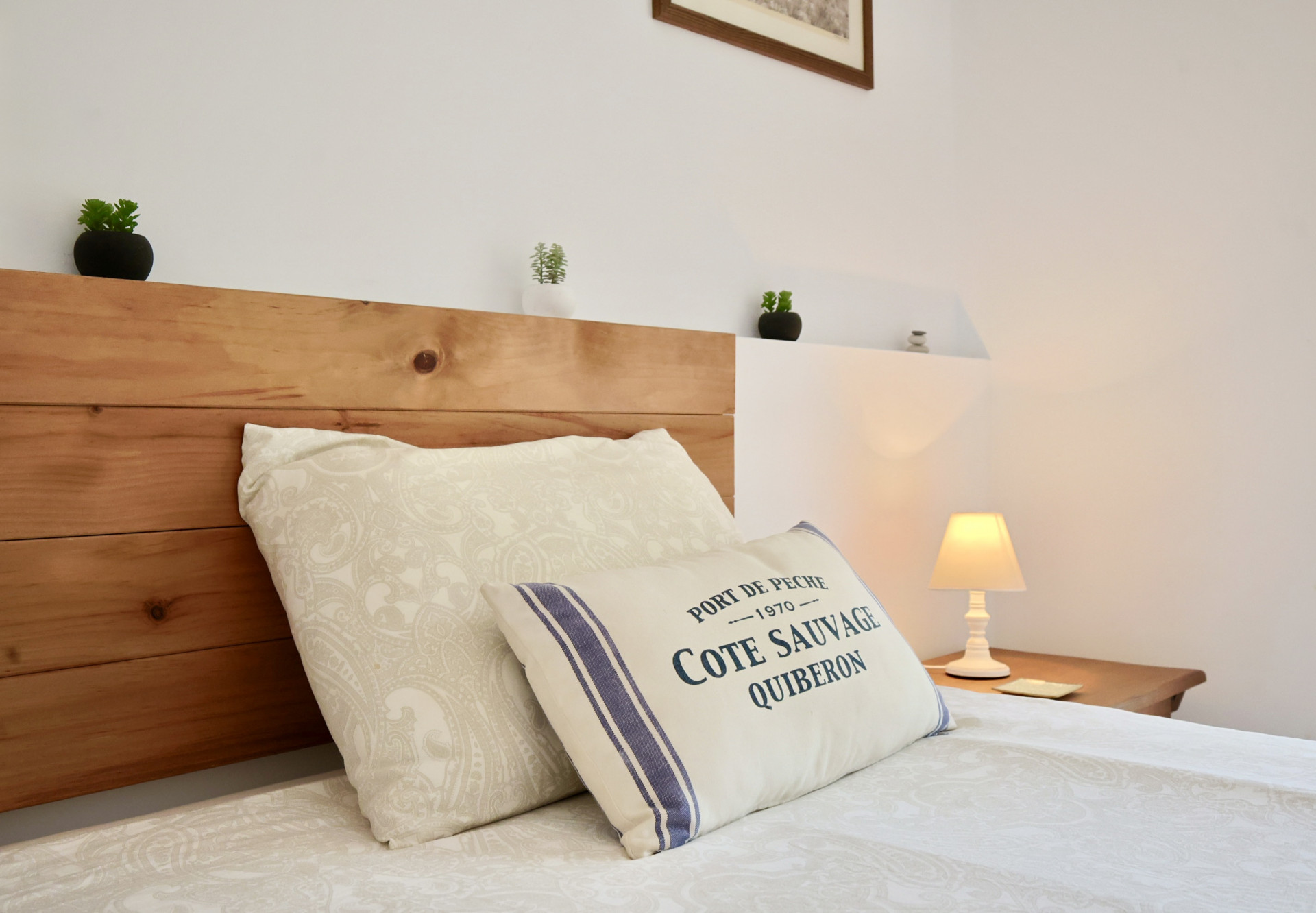 Détail d'une chambre avec tête de lit en bois massif, coussins à motifs marins et lampe de chevet diffusant une lumière tamisée sur la table de nuit.