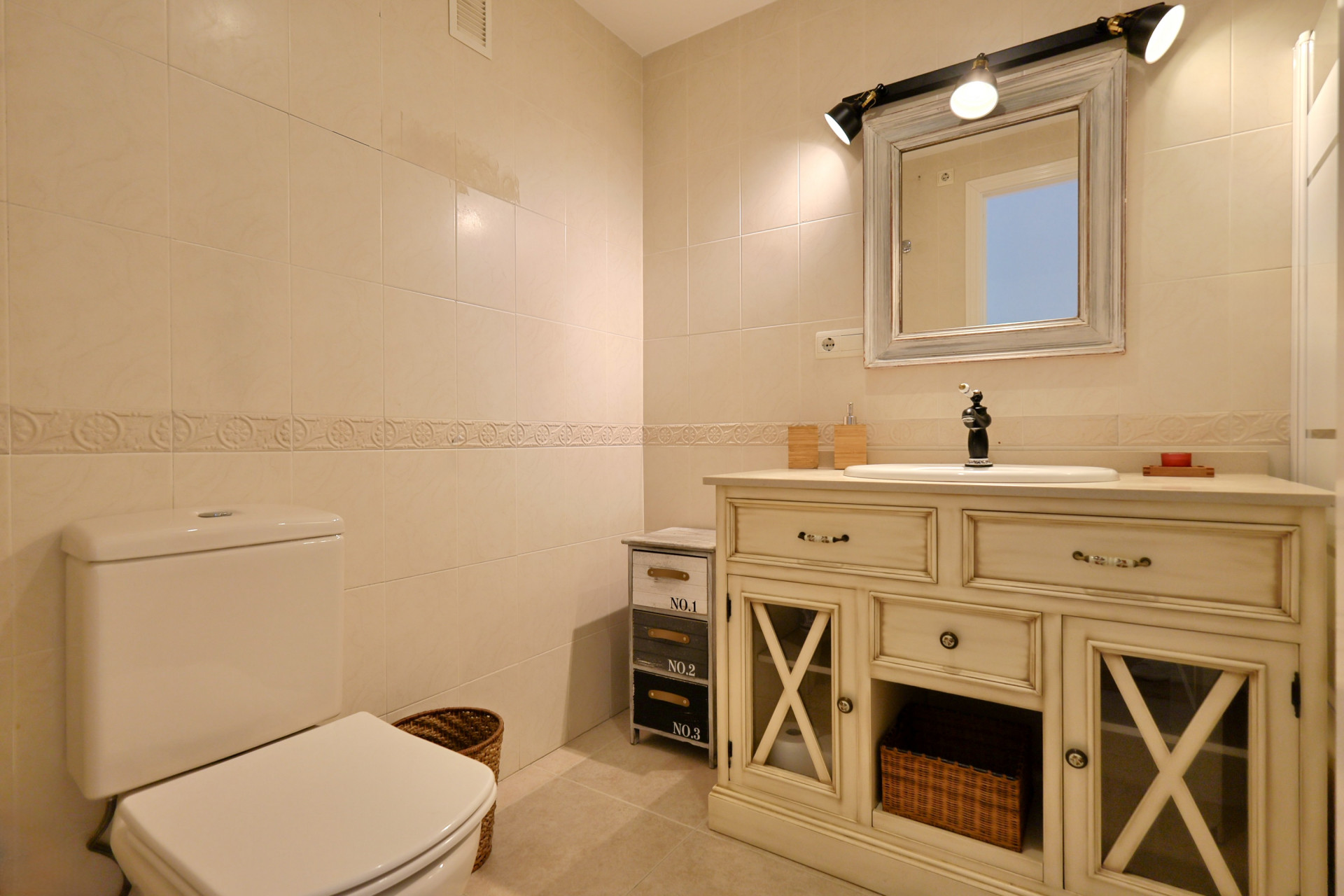 Salle de bain avec meuble vasque de style vintage, portes vitrées à croisillons, robinetterie noire et carrelage mural crème avec frise décorative.