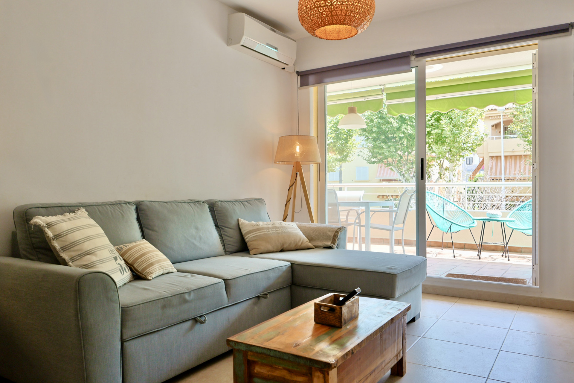 Salon lumineux avec canapé d'angle gris, table en bois rustique et accès à une terrasse meublée équipée de chaises Acapulco turquoise.