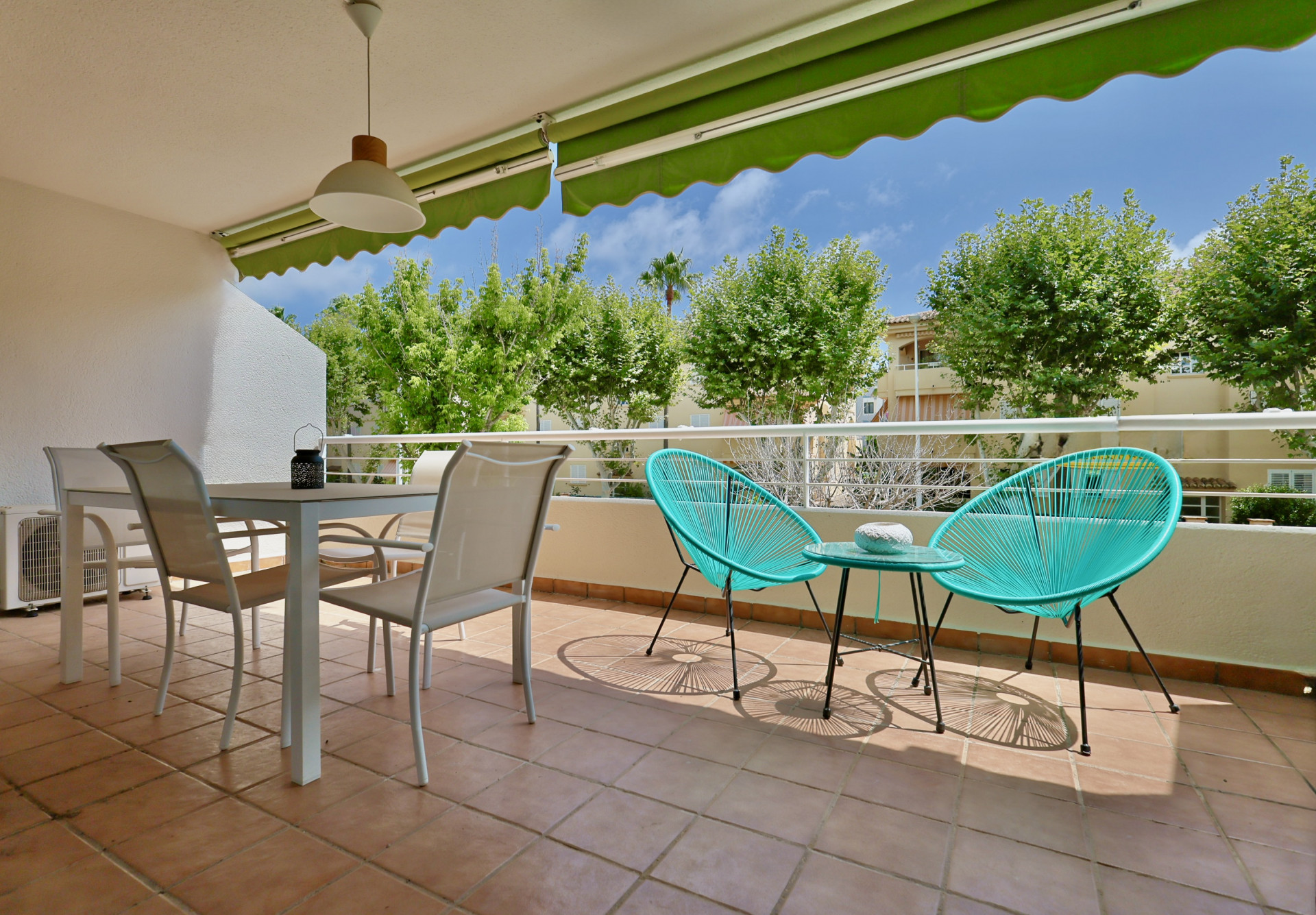 Terrasse extérieure avec carrelage en terre cuite, ensemble de repas blanc et fauteuils Acapulco turquoise sous un store banne vert.