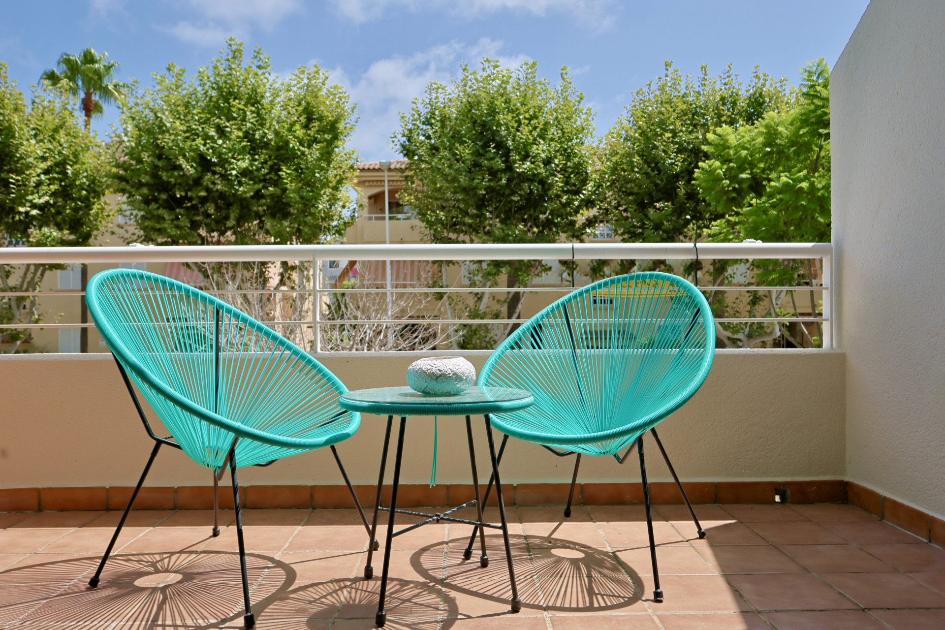 Terrasse privée avec sol en terre cuite, dotée de deux chaises Acapulco turquoise et d'une table assortie offrant une vue sur des arbres verdoyants.