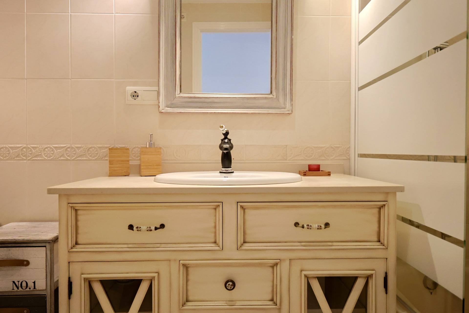 Vanité de salle de bain de style vintage avec meuble crème, robinetterie noire classique et miroir à cadre en bois vieilli sur murs carrelés.