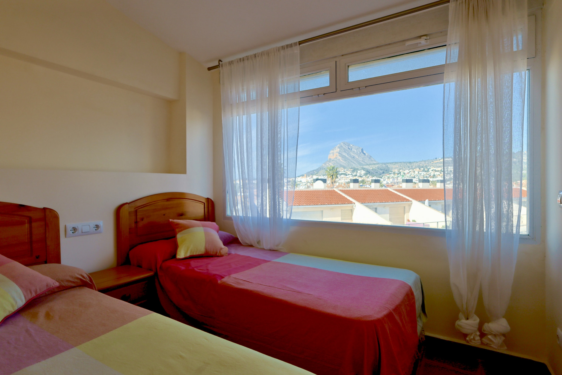Chambre avec deux lits simples, têtes de lit en bois et grande fenêtre offrant une vue sur le mont Montgó et les toits de Jávea.