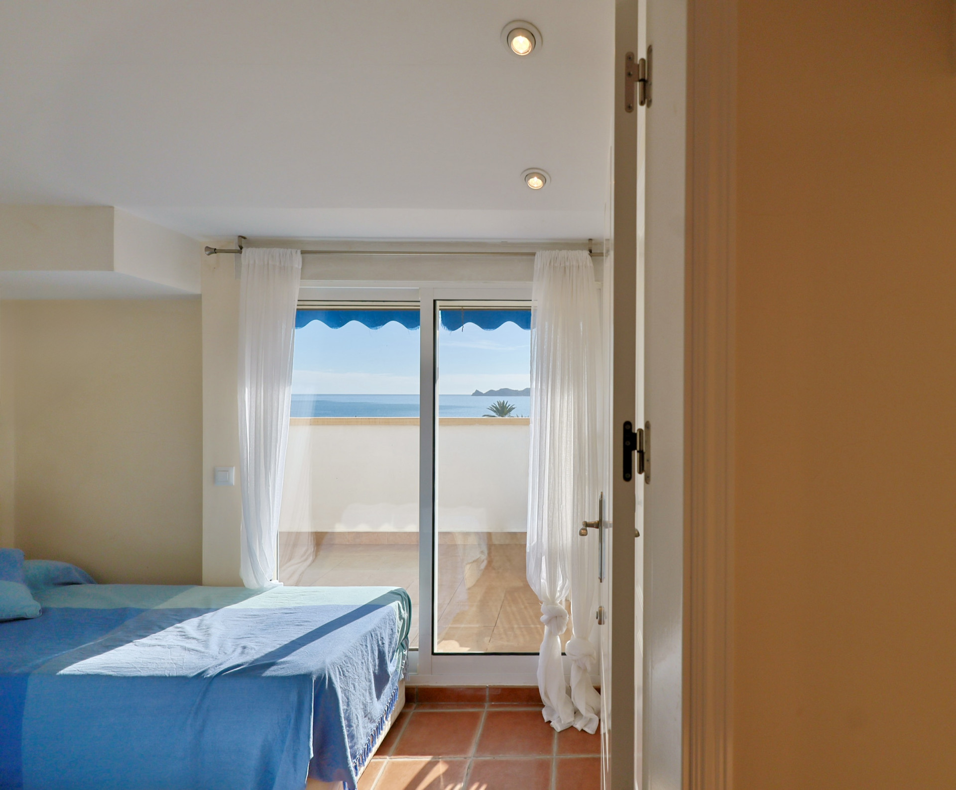 Chambre avec sol en terre cuite et baie vitrée ouvrant sur une terrasse privée offrant une vue sur la mer sous un store banne bleu.