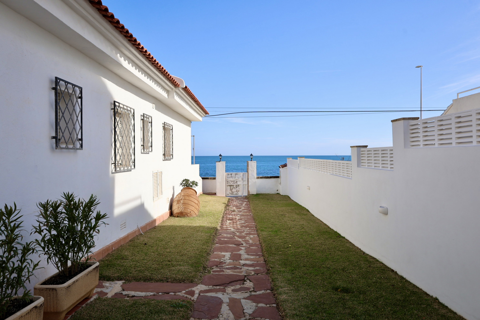 Chemin en pierres naturelles bordé de pelouse menant à un portail blanc avec une vue directe sur la mer Méditerranée à Jávea.