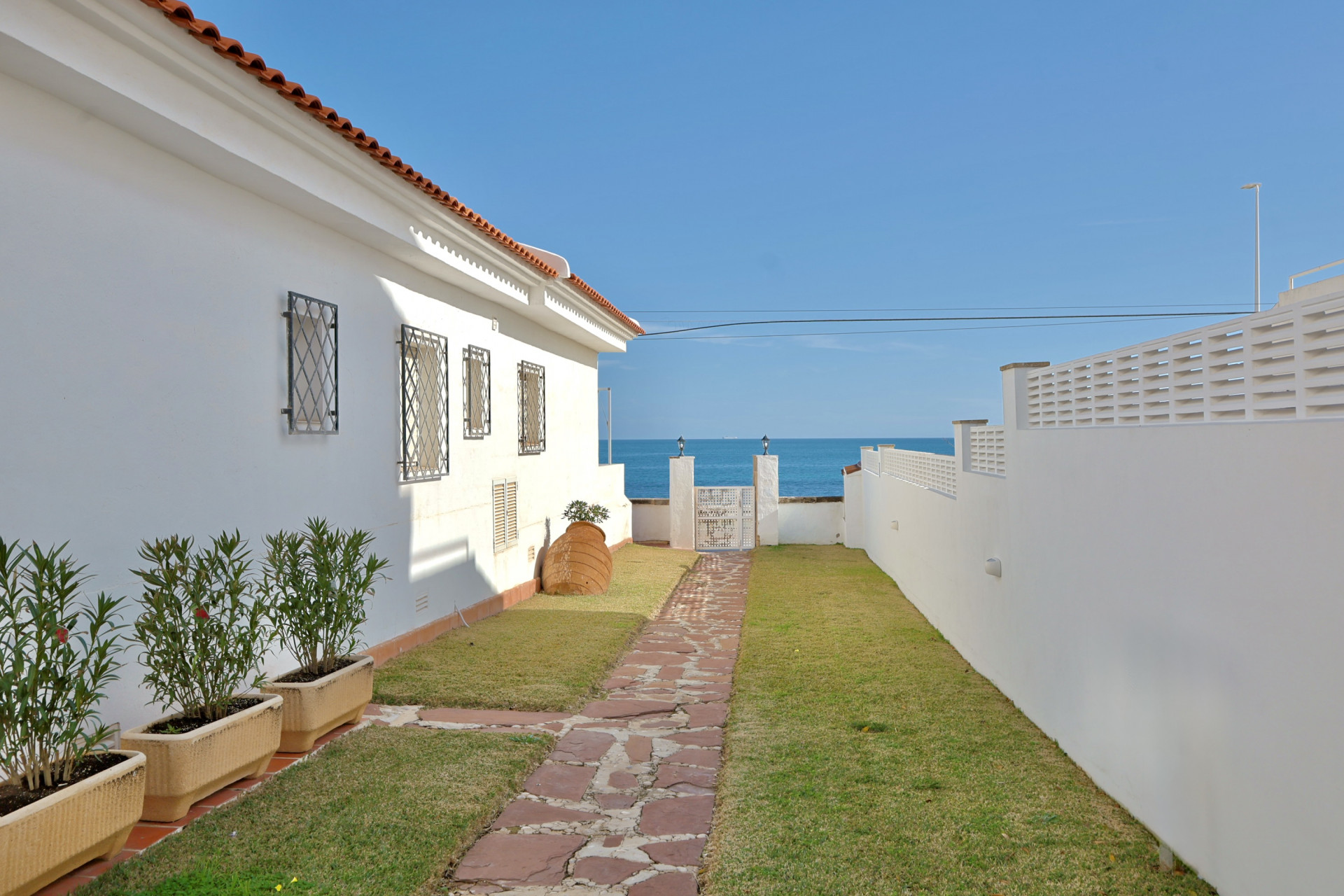 Chemin en pierres naturelles bordé de pelouse menant à un portillon blanc offrant une vue dégagée sur l'horizon et la mer Méditerranée.