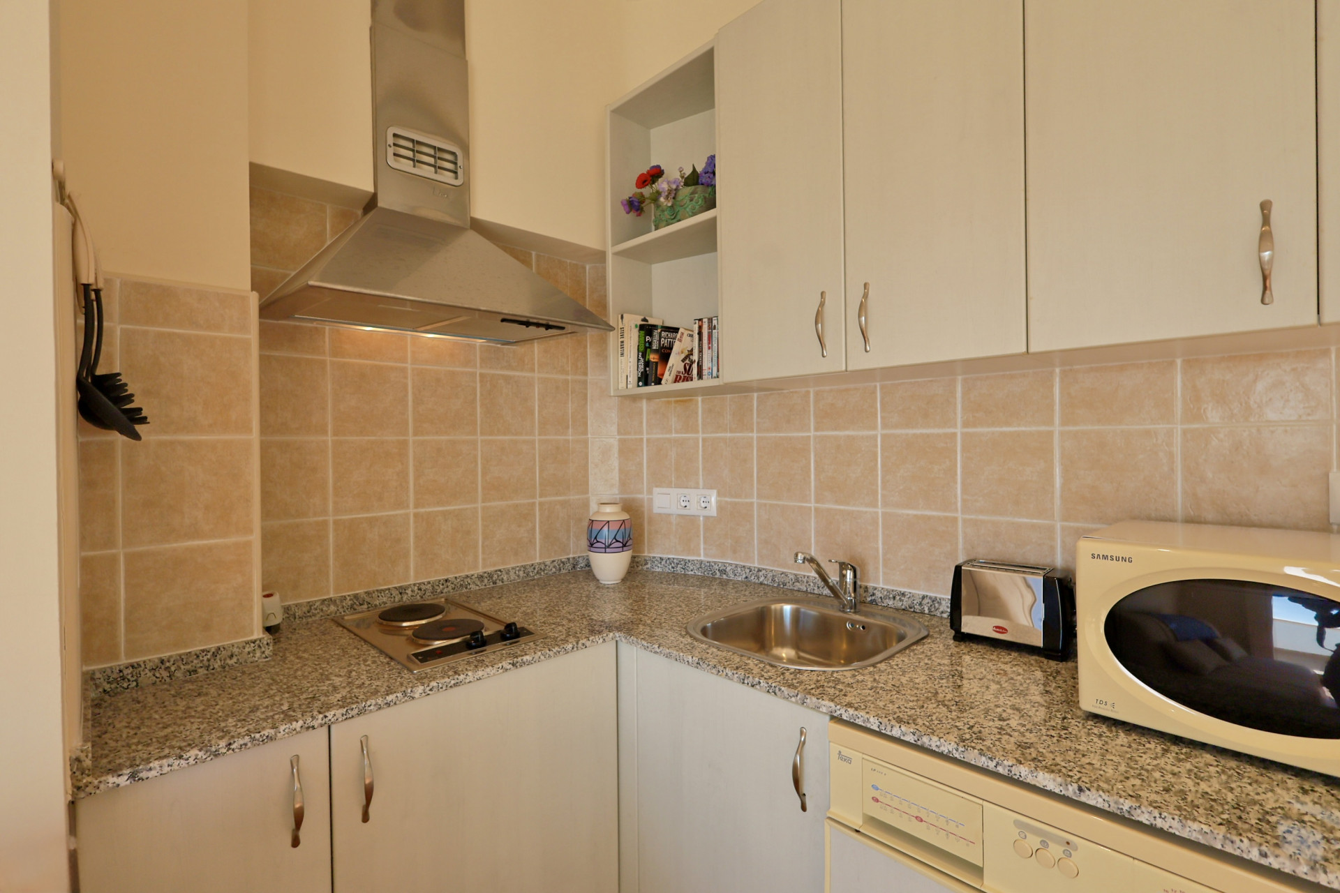 Coin cuisine fonctionnel avec plan de travail en granit moucheté, hotte en inox et carrelage beige. Comprend des rangements blancs et de l'électroménager compact.