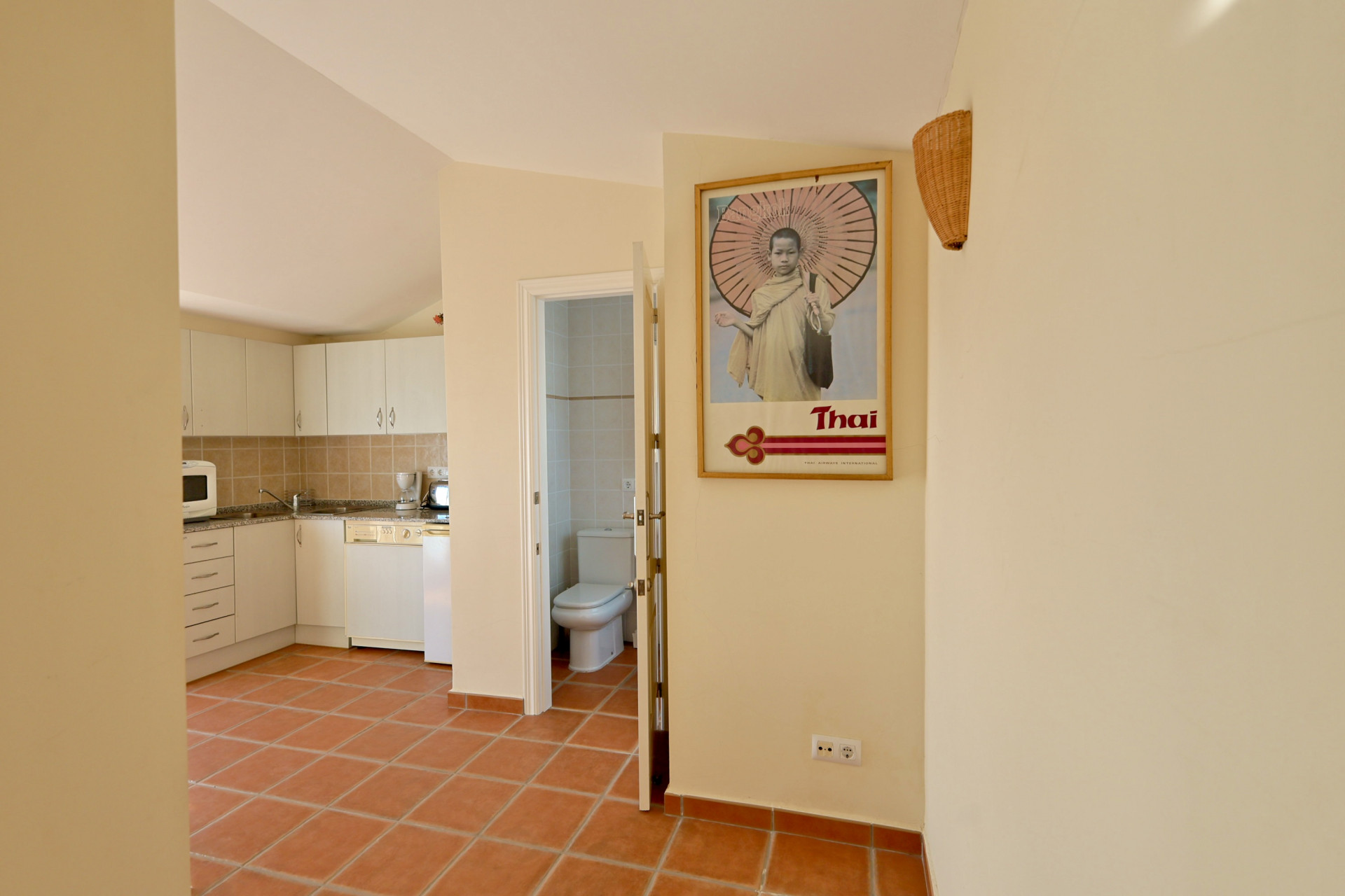 Couloir avec carrelage en terre cuite desservant une cuisine aux façades blanches et une salle de bain carrelée.