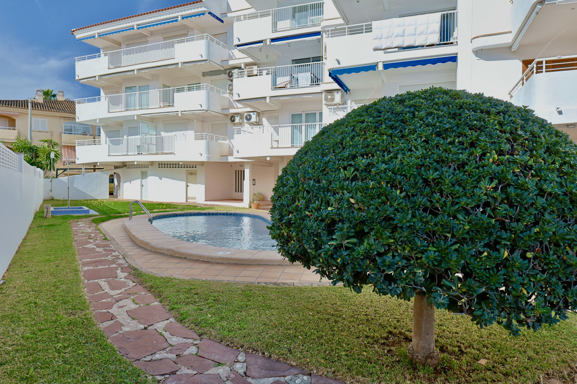 Espace commun avec piscine courbée, pelouse soignée et chemin en dalles de pierre, devant un immeuble blanc équipé de stores bannes bleus.