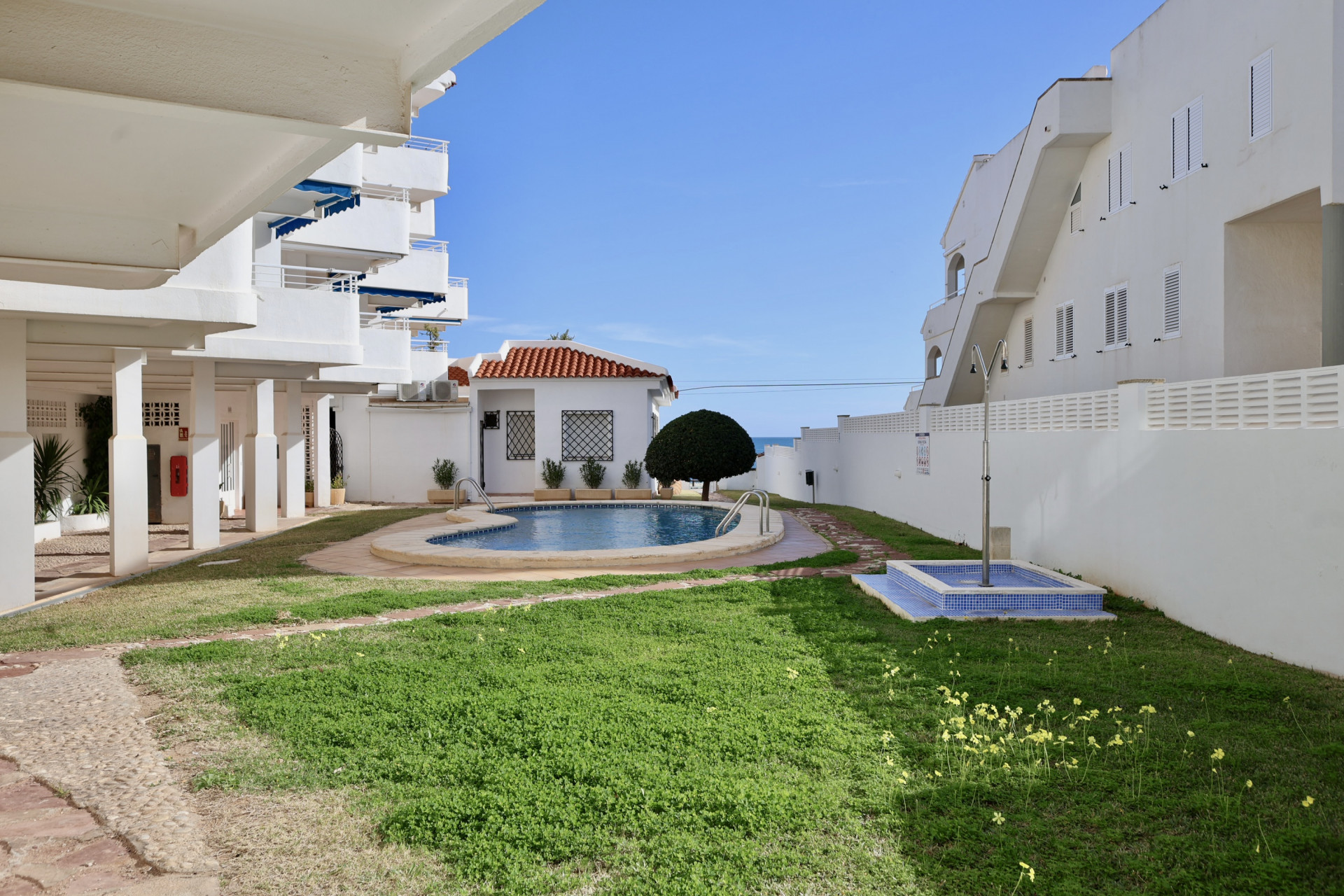 Espace extérieur commun comprenant une piscine en forme de rein, une pelouse et une douche carrelée, entouré de résidences blanches typiques.