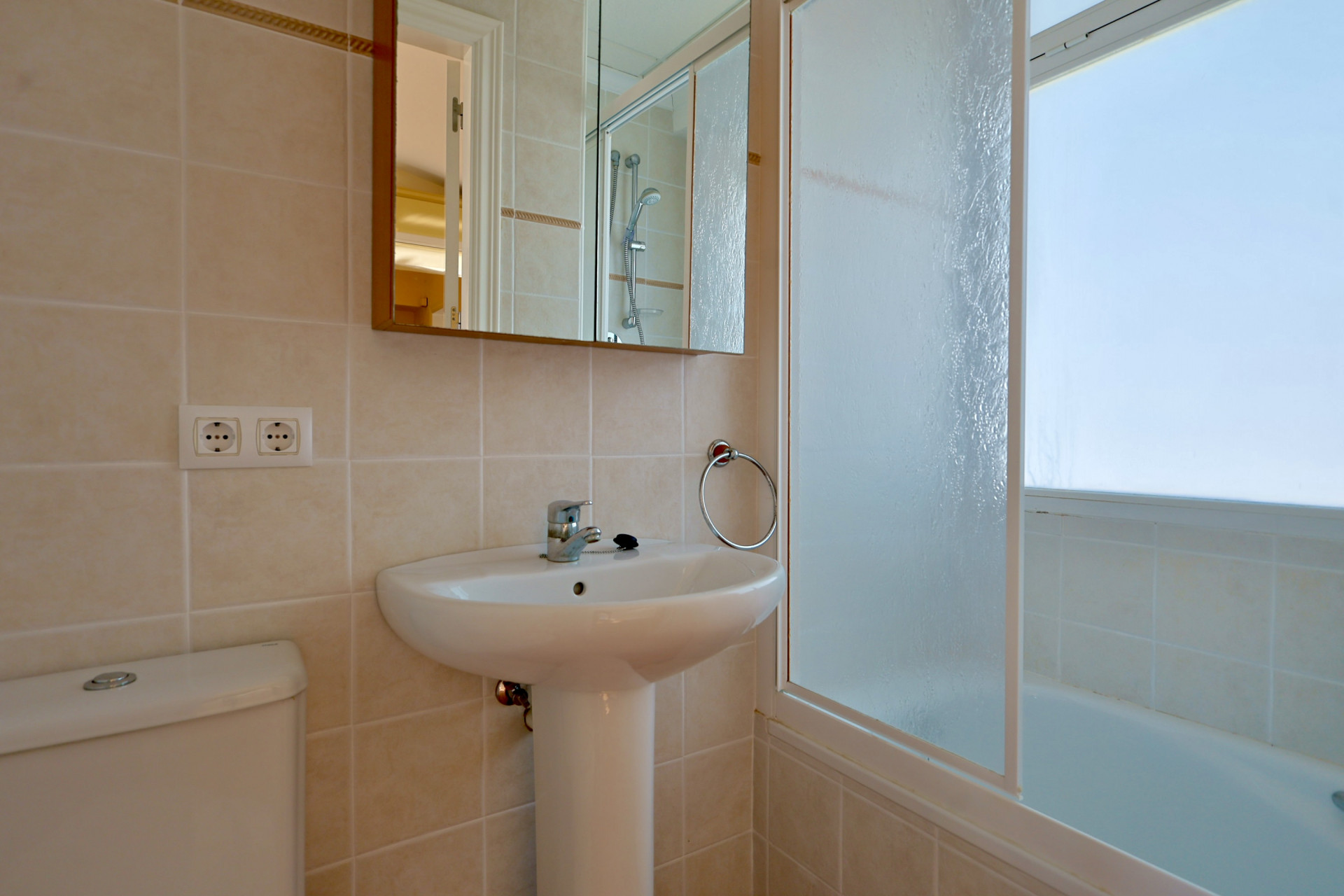 Salle de bain avec lavabo sur pied, armoire de toilette à miroir et baignoire équipée d'un pare-douche en verre dépoli. Carrelage mural beige.
