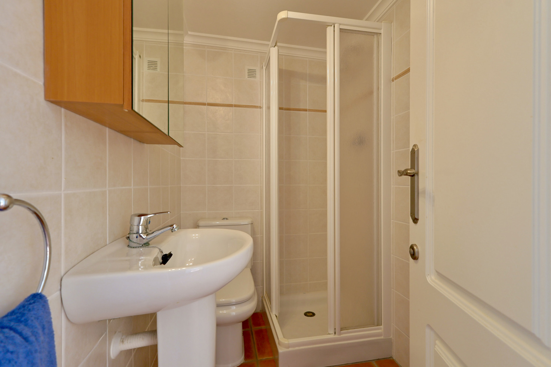 Salle de bain fonctionnelle équipée d'un lavabo sur colonne, d'une douche d'angle et de carrelage mural beige avec frise décorative.