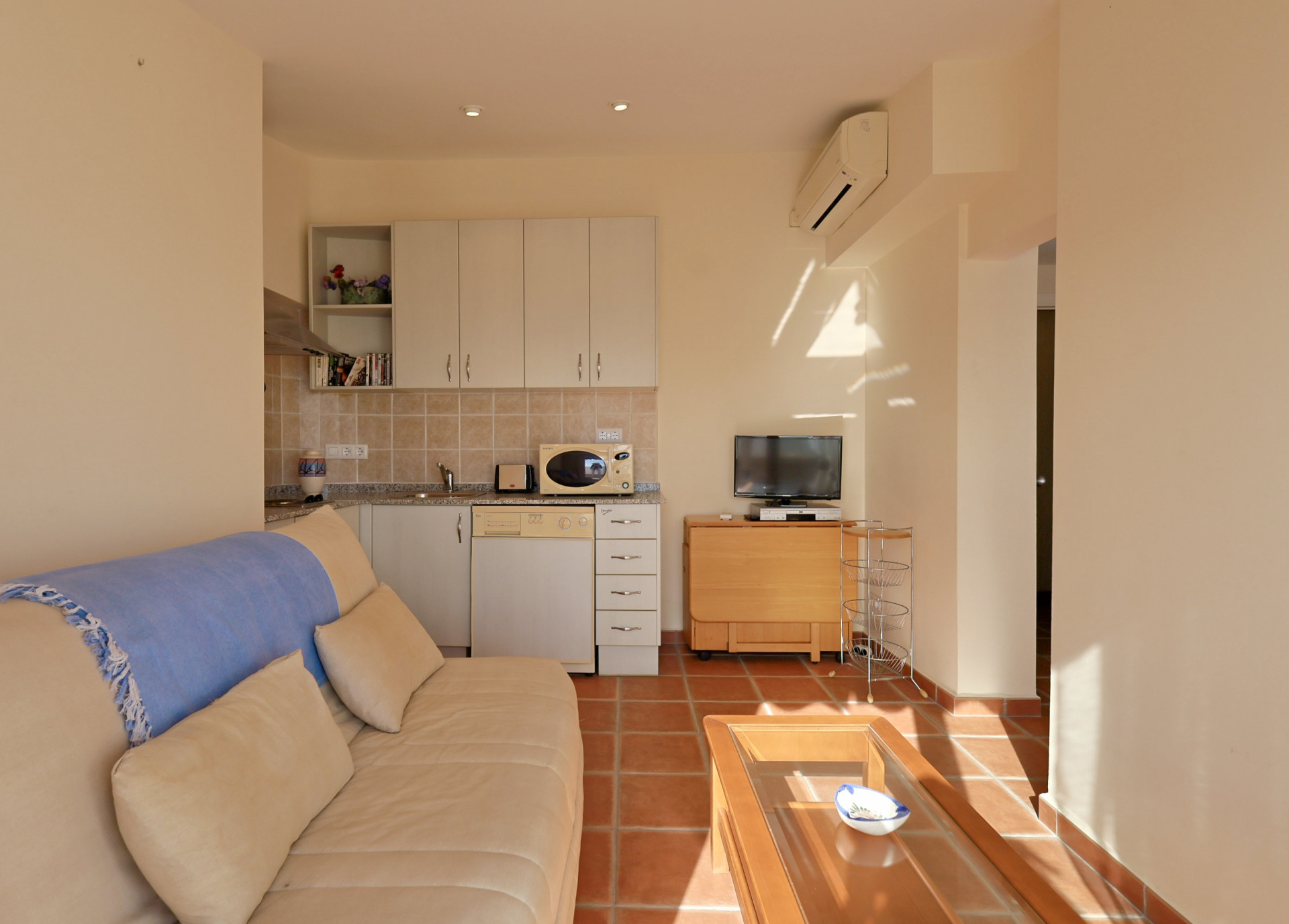 Salon avec sol en terre cuite, kitchenette intégrée aux meubles blancs, canapé crème et climatisation murale réversible.