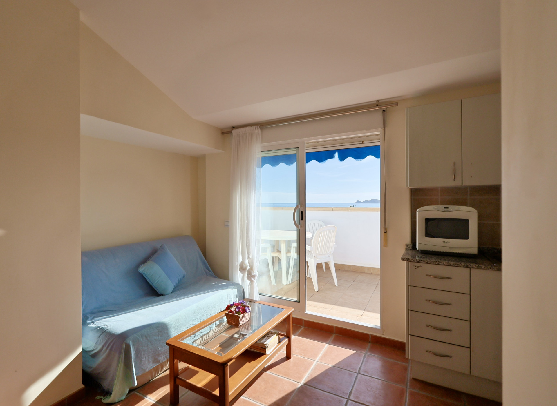 Salon lumineux avec sol en terre cuite, canapé bleu et accès à une terrasse privée offrant une vue sur la mer sous un store bleu.