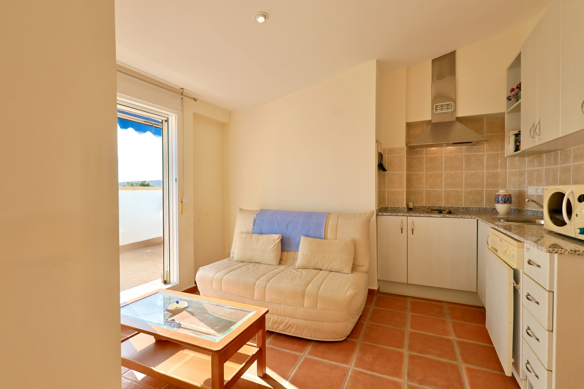 Salon ouvert avec carrelage en terre cuite, kitchenette avec plans de travail en granit et accès direct à une terrasse ensoleillée par une porte vitrée.