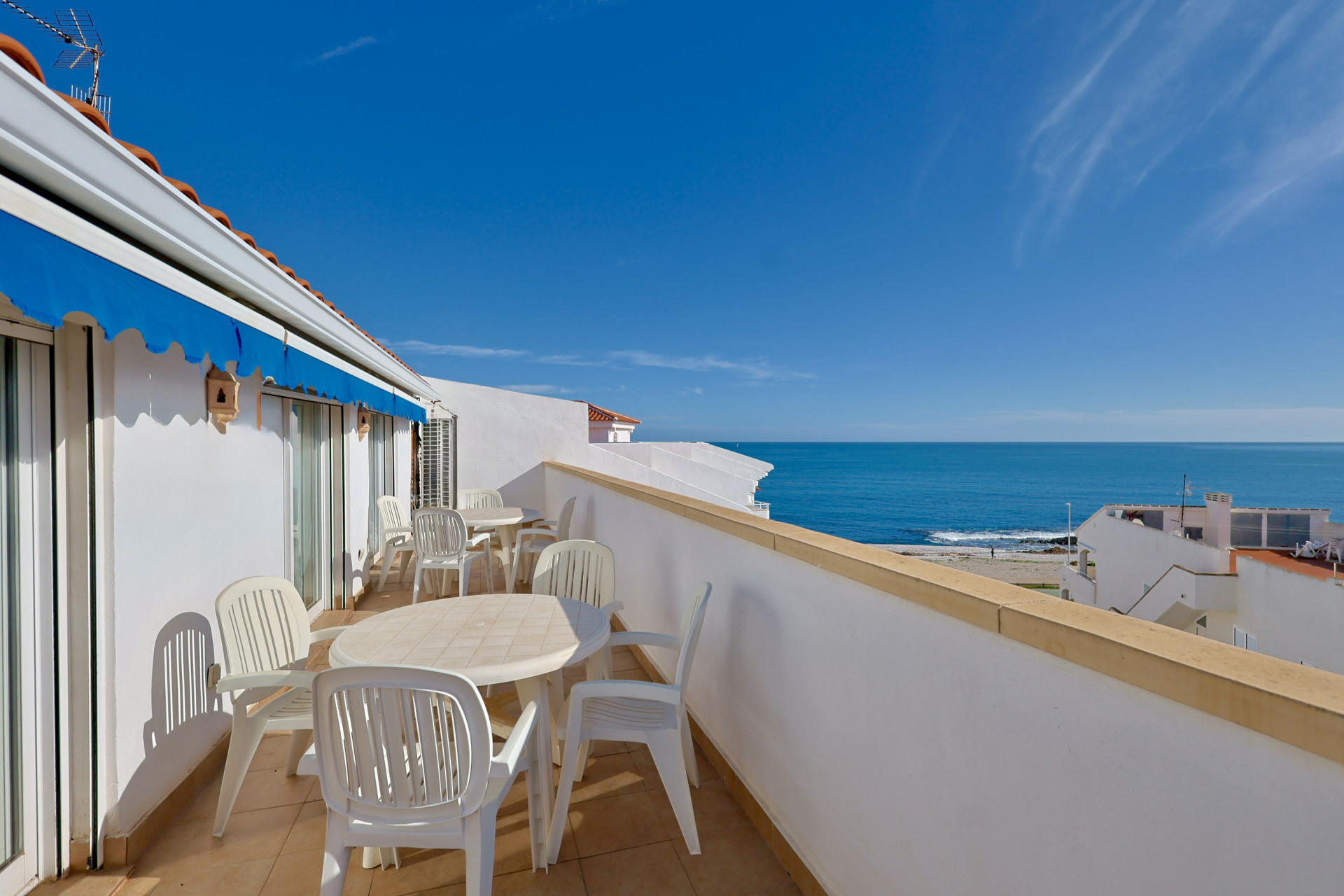 Terrasse en première ligne avec carrelage en grès, mobilier d'extérieur blanc et store bleu escamotable offrant une vue dégagée sur la mer.