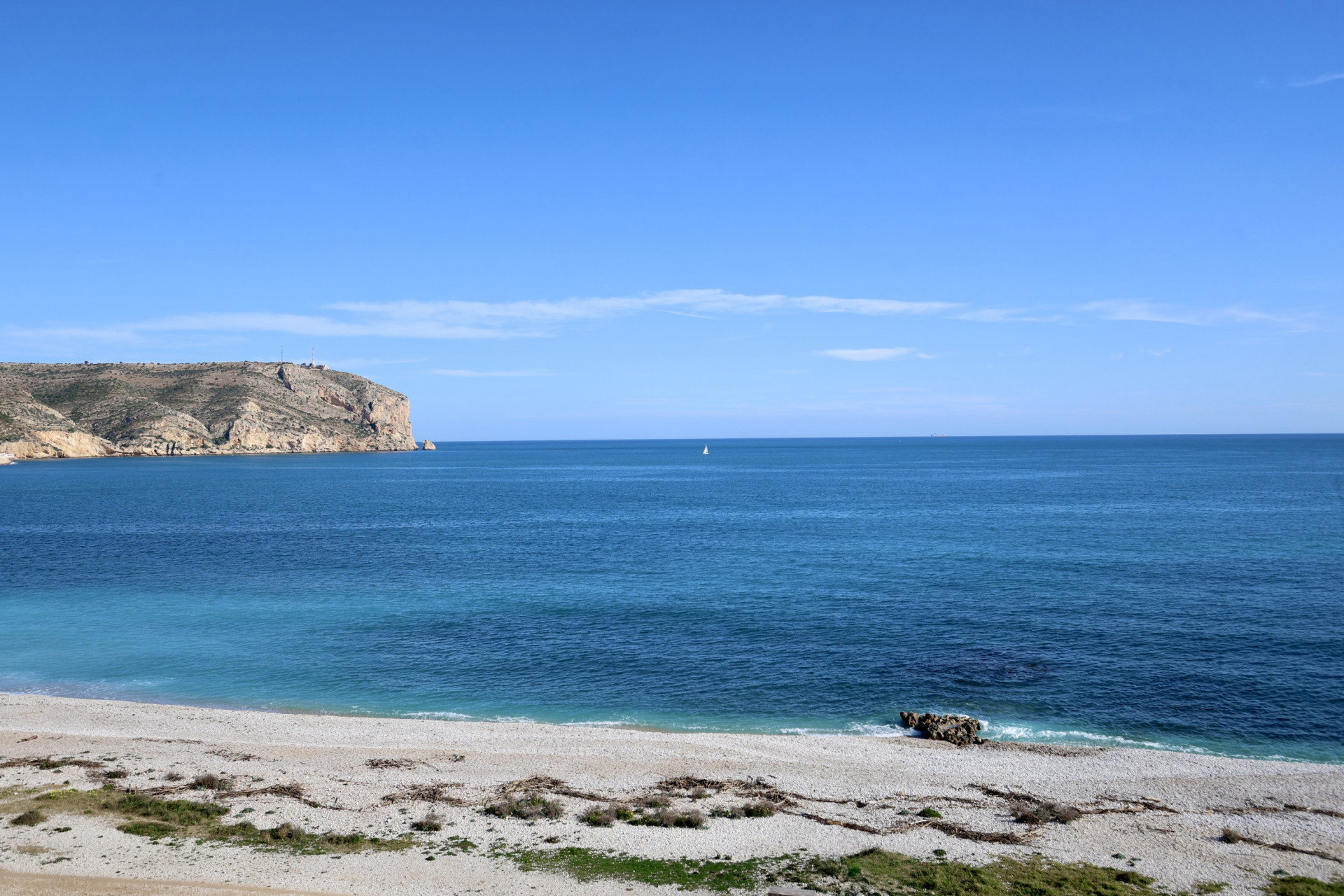 Vue frontale sur la mer Méditerranée et le cap San Antonio depuis la plage de galets à Jávea, sous un ciel bleu dégagé.