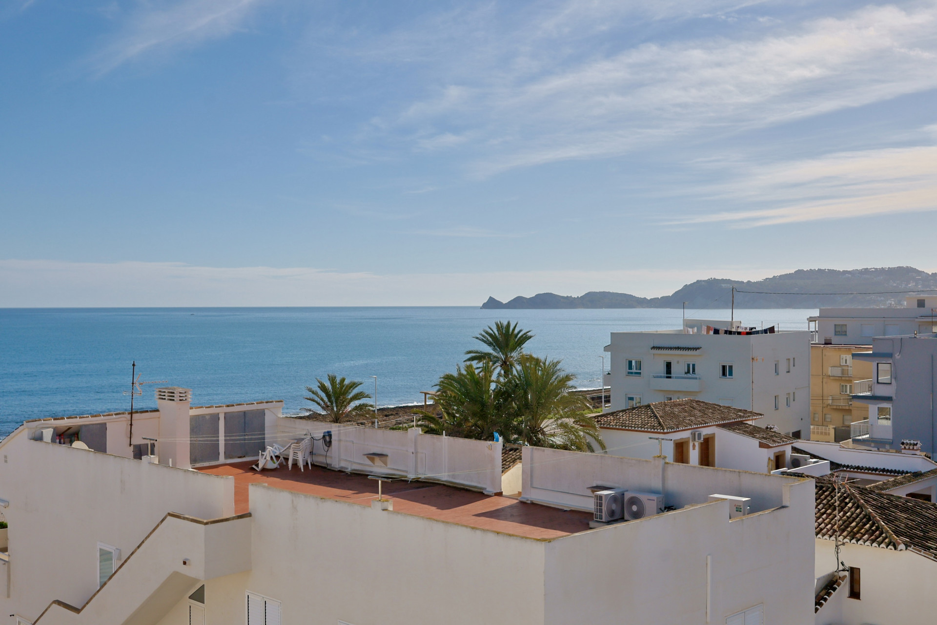 Vue panoramique depuis le toit-terrasse sur la mer Méditerranée, avec des bâtiments blancs, des palmiers et les montagnes côtières à l'horizon.