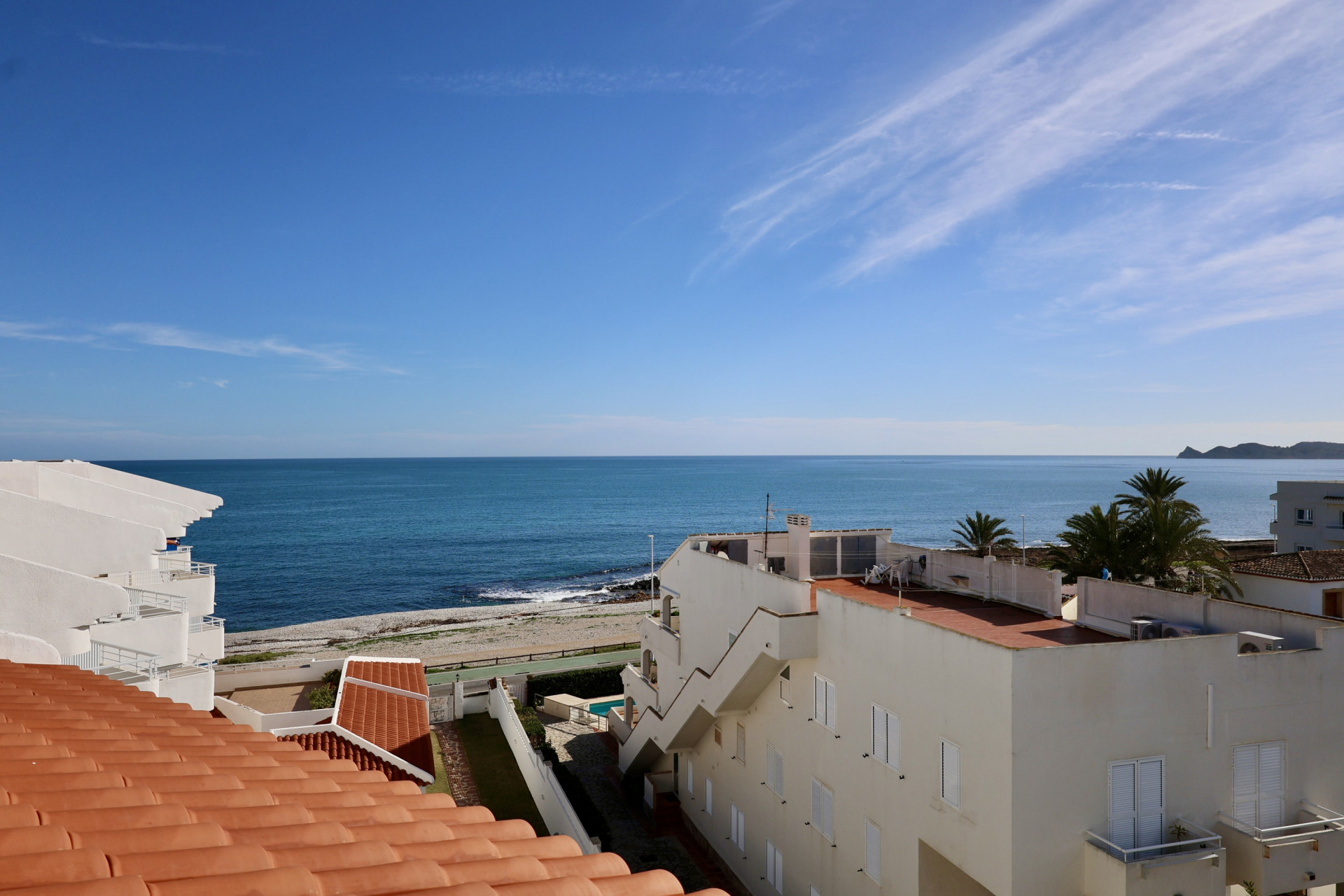 Vue surélevée sur les toits en terre cuite vers la mer Méditerranée et le rivage de galets, avec des bâtiments côtiers blancs sous un ciel bleu clair à Jávea.