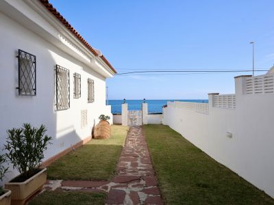 Allée de jardin extérieure en dalles de pierre menant à un portail offrant une vue directe sur la mer Méditerranée. La maison blanche arbore des grilles en fer forgé.