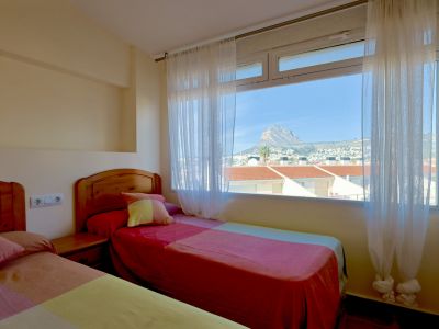 Chambre lumineuse avec deux lits simples à têtes de lit en bois, dotée d'une grande fenêtre offrant une vue dégagée sur la montagne Montgó et les toits.