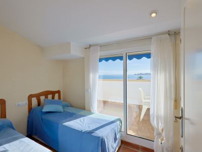 Chambre lumineuse avec deux lits simples et accès direct à un balcon privé offrant une vue panoramique sur la mer et la côte. Sol carrelé en terre cuite et lumière naturelle abondante.