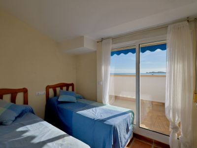 Chambre lumineuse avec deux lits simples et têtes de lit en bois. Accès à un balcon carrelé de terre cuite, offrant une vue sur la mer avec un auvent bleu.