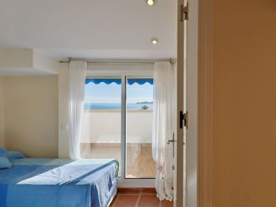 Chambre lumineuse avec lit habillé de bleu et accès direct à une terrasse offrant une vue panoramique sur la mer Méditerranée. Sol en terre cuite et rideaux blancs.