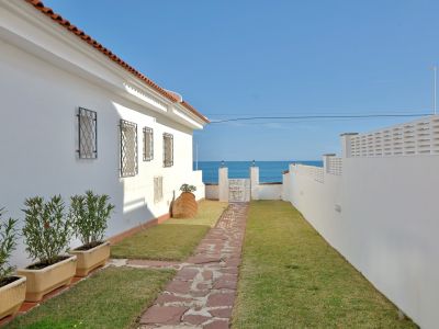 Chemin de pierres menant à un portail blanc, révélant une vue directe sur la mer sous un ciel bleu. Un bâtiment blanc avec un toit en tuiles et des fenêtres grillagées borde la pelouse.