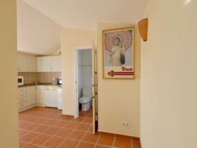 Couloir avec carrelage en terre cuite menant à une cuisine ouverte aux armoires blanches et une salle de bain visible. Une affiche 'Thai' décore le mur.