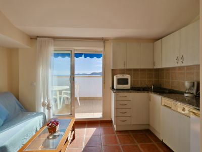 Espace de vie ouvert avec cuisine compacte aux armoires blanches et sol en terre cuite. Porte coulissante vitrée menant à un balcon avec vue sur la mer et store bleu.