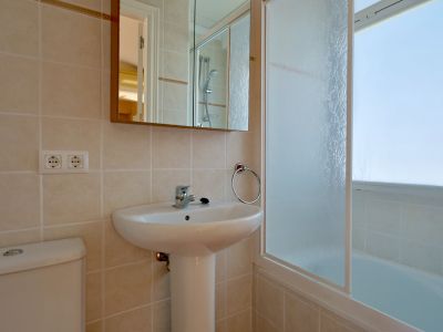 Salle de bain moderne avec lavabo sur pied blanc, toilettes et murs carrelés clairs. Un miroir-armoire est au-dessus du lavabo, jouxtant une baignoire avec écran en verre dépoli.
