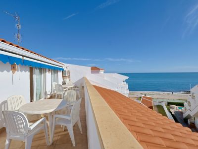 Terrasse extérieure aménagée avec mobilier blanc, un store bleu et une vue dégagée sur la mer Méditerranée et la côte.