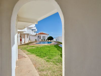Vue à travers une arche blanche encadrant une piscine commune avec pelouse, un bâtiment au toit de tuiles et la mer Méditerranée au loin sous un ciel bleu clair.