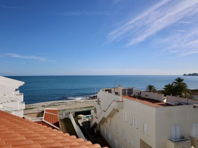 Vue de toit avec tuiles en terre cuite, présentant un panorama sur la mer Méditerranée et une plage de galets. Bâtiments côtiers blancs et un cap lointain sous un ciel bleu clair.