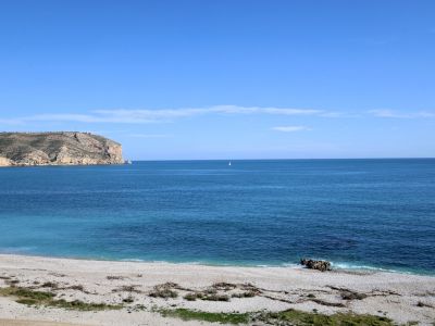 Vue panoramique d'une plage de galets avec les eaux turquoise de la Méditerranée, un voilier solitaire à l'horizon et une falaise rocheuse à gauche.