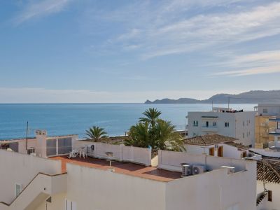 Vue panoramique sur la mer Méditerranée avec la côte de Jávea à l'horizon, toits d'architecture locale et palmiers sous un ciel clair.