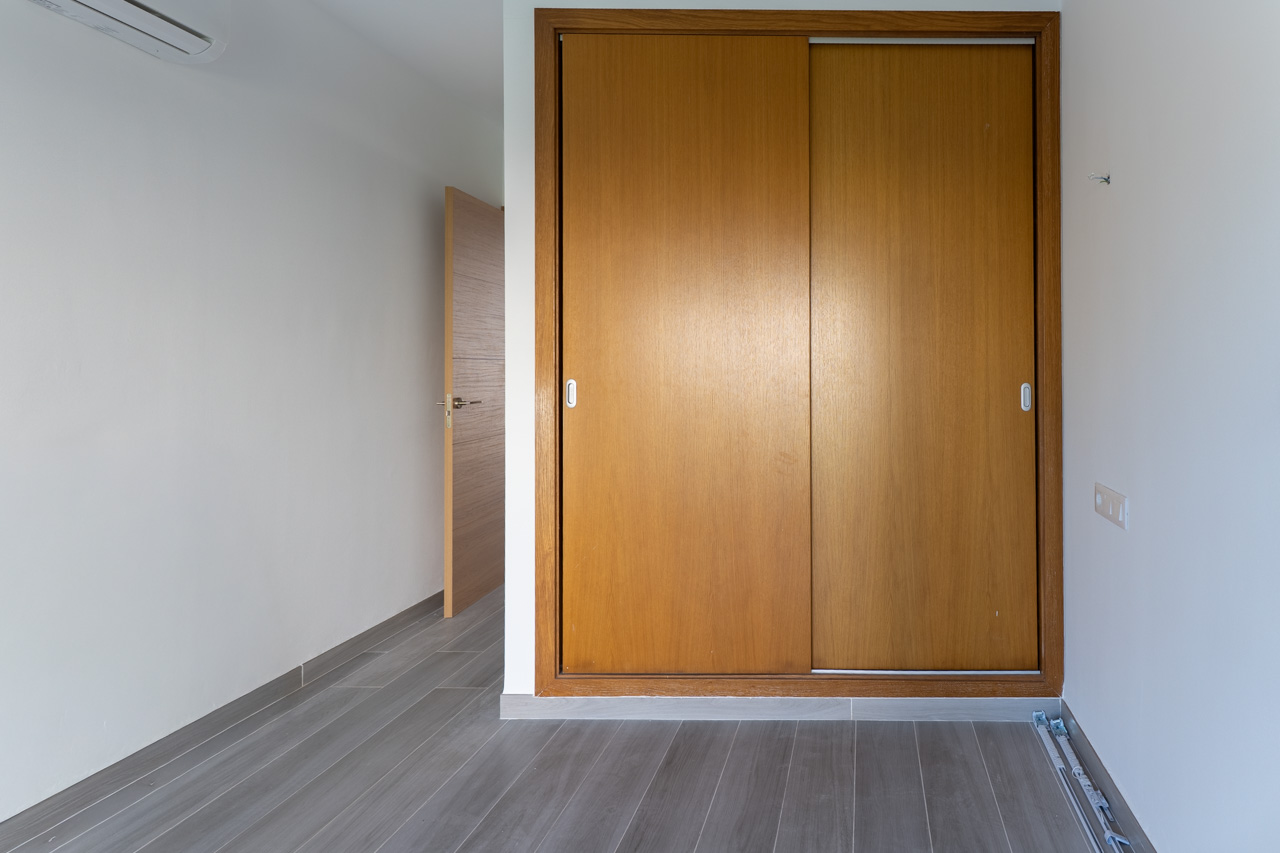 Armoire encastrée avec portes coulissantes en bois, sol en grès cérame gris imitation parquet et murs blancs. Comprend une unité de climatisation murale.