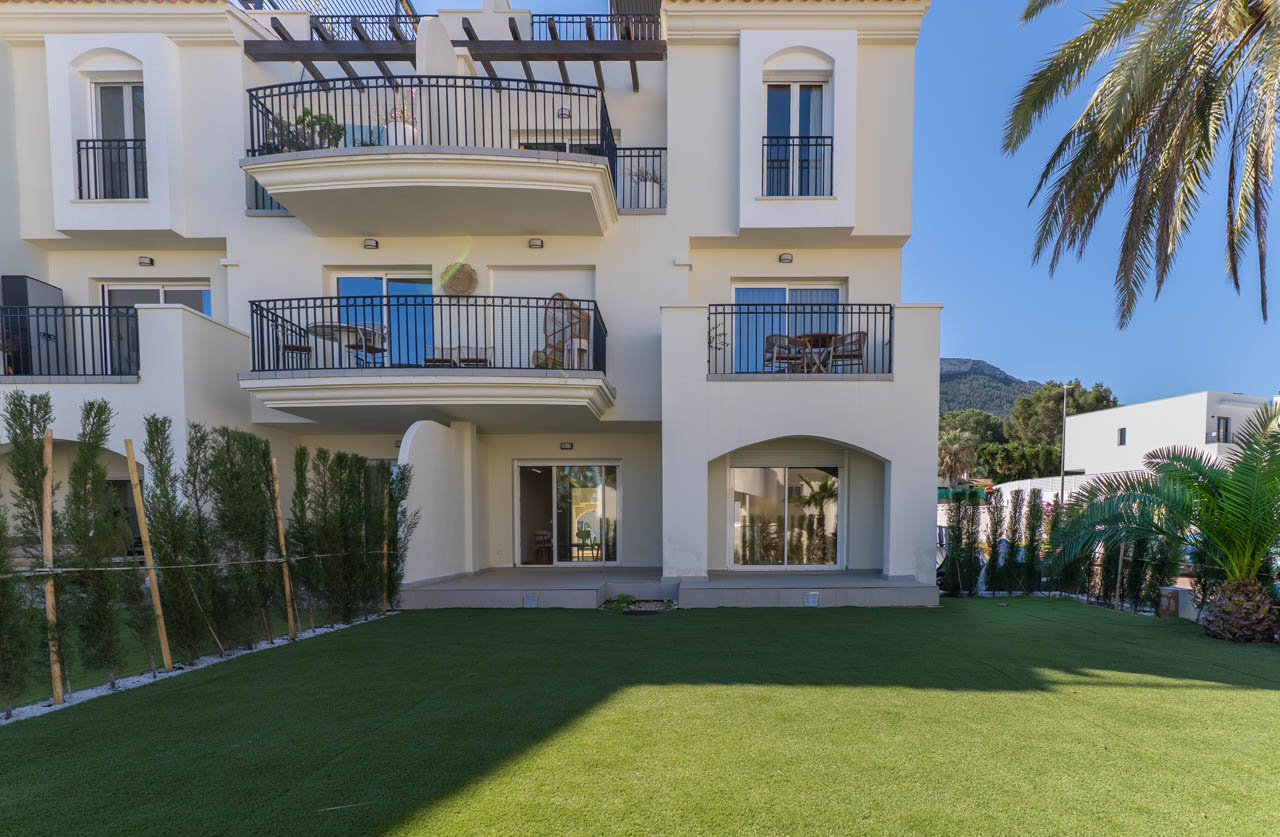 Façade blanche avec balcons superposés en fer forgé noir et un jardin privé au rez-de-chaussée avec gazon artificiel, bordé de cyprès et de palmiers.