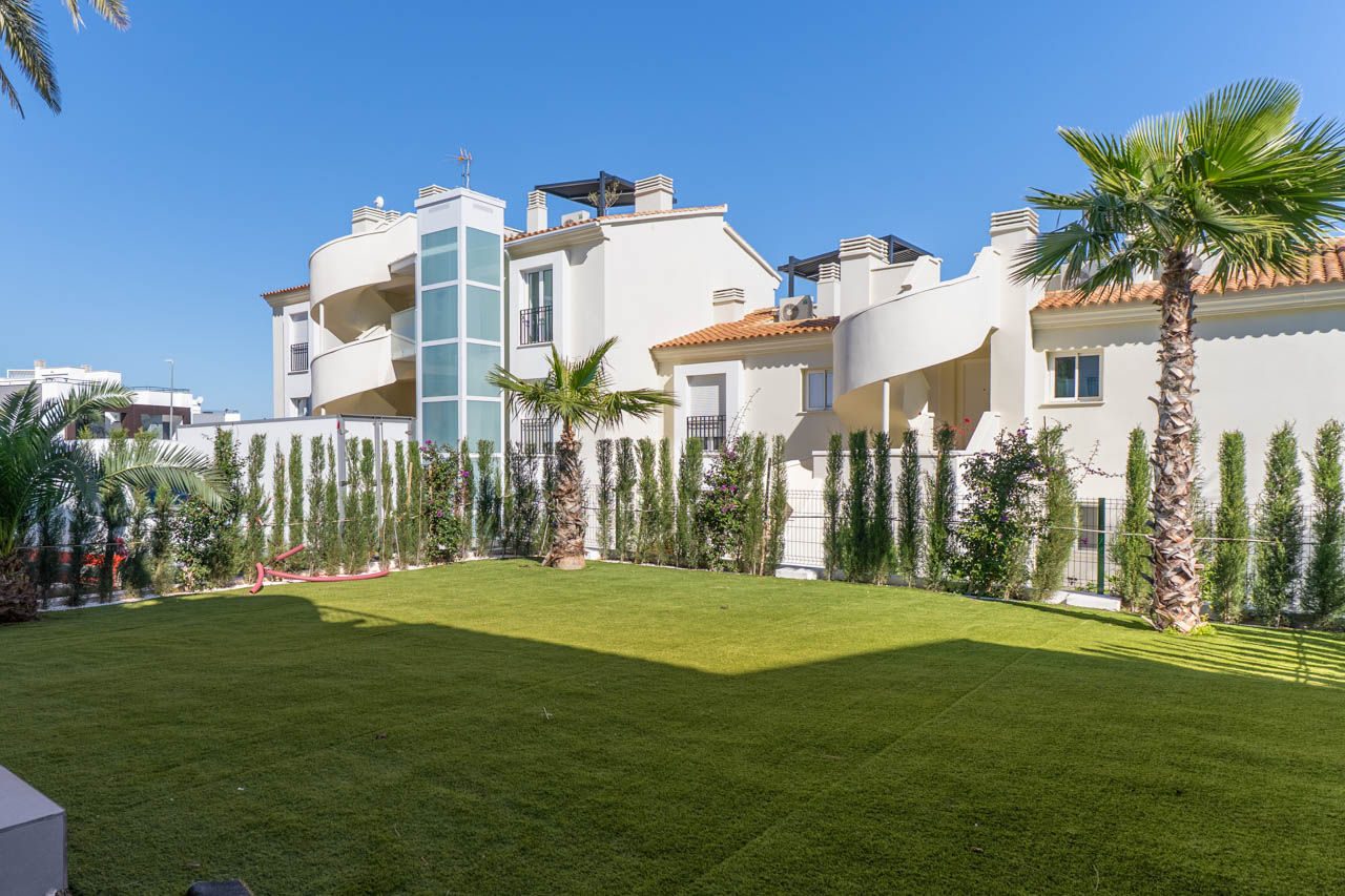 Jardin privé avec gazon synthétique, palmiers et haie de cyprès. La façade blanche se distingue par ses escaliers en colimaçon et son ascenseur vitré moderne.
